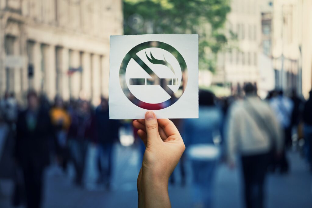 Hand holding a cutout of a 'no-smoking' emblem in front of a crowded street